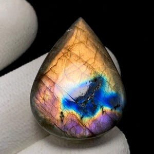 May include: A teardrop-shaped labradorite gemstone with a polished surface. The stone displays a vibrant play of color, with hues of blue, purple, and gold. The gemstone is set against a dark background, highlighting its iridescent qualities.