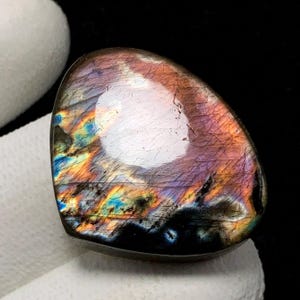 May include: A polished, iridescent labradorite gemstone. The stone displays a mix of colors, including purple, orange, blue, and green, with a prominent white area. The gemstone has a smooth, rounded shape and a dark base.