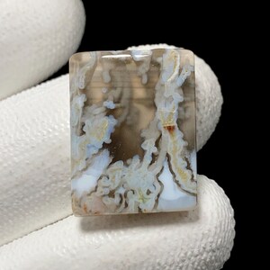 May include: A rectangular gemstone with a unique pattern of brown, white, and blue swirls. The polished surface of the stone has a translucent quality, revealing intricate details. The stone is held by a gloved hand, highlighting its natural beauty.