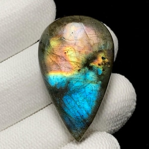 May include: A teardrop-shaped labradorite gemstone with a polished surface. The stone displays a vibrant play of color, with iridescent flashes of blue, orange, and yellow. The gemstone is held against a white fabric background.