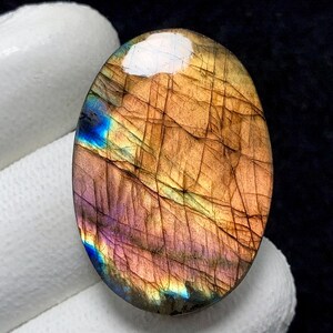 May include: Oval-shaped labradorite gemstone with a polished surface. The stone displays a mix of colors, including brown, purple, blue, and gold, with visible internal lines. The gemstone is held by a gloved hand.