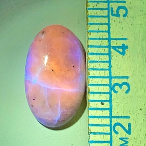 May include: Oval-shaped gemstone with a peach and lavender color gradient. The stone has internal veining and a glossy surface. A ruler is positioned next to the stone, with markings in inches.