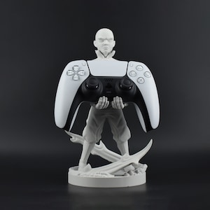May include: A white and black PlayStation 5 controller held by a gray figurine. The figurine is standing on a round base with a detailed design. The controller is the focal point, with its buttons and design clearly visible.