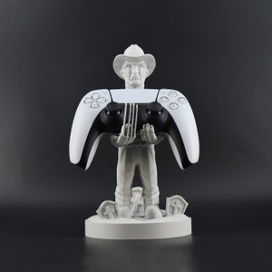 Puede incluir: Una figura blanca impresa en 3D de una persona con un sombrero de vaquero sosteniendo un mando de PlayStation 5 blanco y negro. La figura está de pie sobre una base blanca con un tema de cementerio.