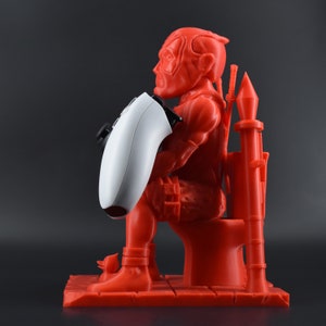 Peut inclure: Une figurine rouge en 3D imprim&eacute;e d'un personnage assis sur des toilettes tenant une manette de jeu vid&eacute;o blanche. La figurine tient la manette devant elle avec sa main gauche. La figurine porte une tenue rouge et noire. Les toilettes sont &eacute;galement rouges et ont un si&egrave;ge blanc. La figurine est assise sur une base rouge.