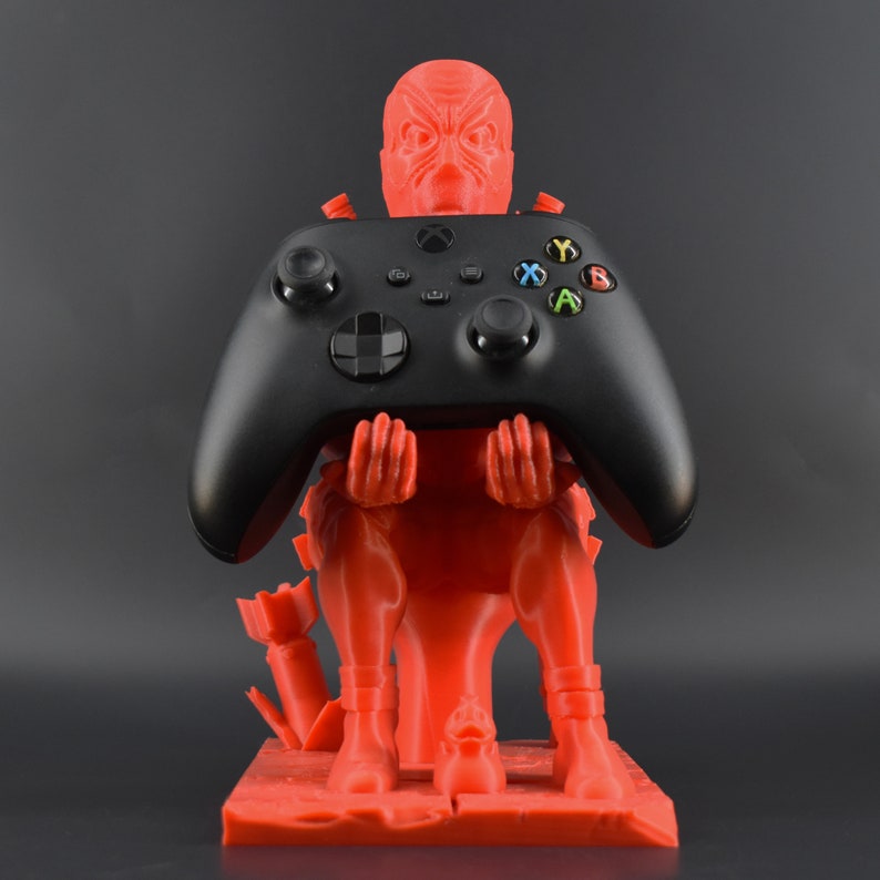 Peut inclure: Une figurine rouge en 3D imprim&eacute;e d'une cr&eacute;ature tenant une manette Xbox noire. La figurine est assise sur une base rouge.