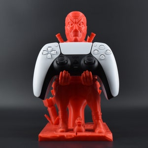 Peut inclure: Une figurine rouge en 3D imprim&eacute;e d'un personnage tenant une manette DualSense PlayStation 5 blanche. La figurine est debout sur une base rouge avec une petite figurine de canard jaune.
