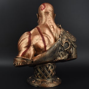 Puede incluir: Un busto dorado de una figura calva y musculosa con marcas rojas. La figura lleva un casco con barba y est&aacute; de pie sobre una base dorada.