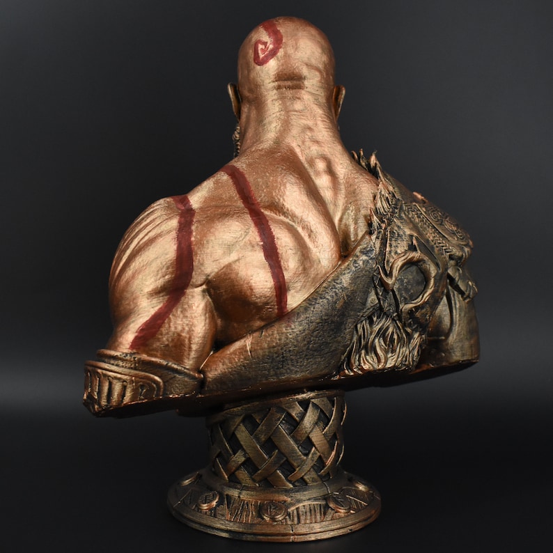 Puede incluir: Un busto dorado de una figura calva y musculosa con marcas rojas. La figura lleva un casco con barba y est&aacute; de pie sobre una base dorada.