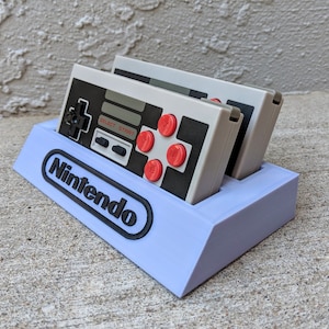 Op de afbeelding: Een paarse plastic standaard met het woord "Nintendo" erop gedrukt. Twee grijs-zwarte Nintendo Entertainment System-controllers staan in de standaard.