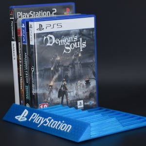 Può includere: Un supporto per giochi PlayStation in plastica blu con il logo PlayStation sul davanti. Il supporto contiene cinque custodie di videogiochi, tra cui Demon's Souls, Assassin's Creed, Uncharted 2: Among Thieves e Tekken 5.