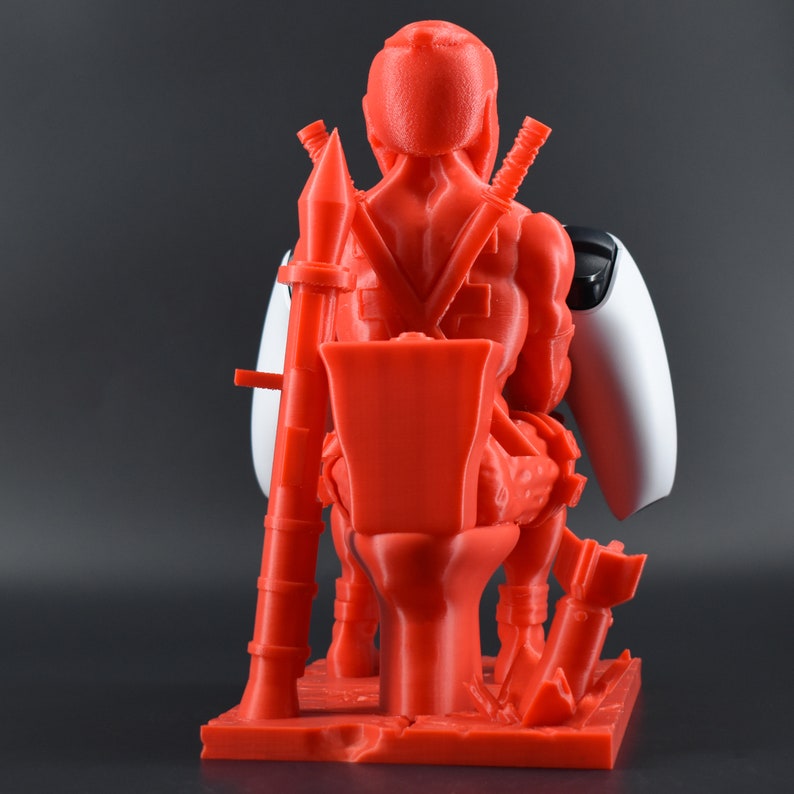 Peut inclure: Une figurine rouge en 3D d'un super-h&eacute;ros tenant une manette de jeu vid&eacute;o blanche. La figurine est assise sur une base rouge en 3D avec un pistolet rouge en 3D et une &eacute;p&eacute;e rouge en 3D.