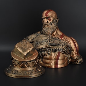 Puede incluir: Un busto de bronce de un guerrero con barba y marcas rojas en la cara. El busto lleva una armadura detallada con un dise&ntilde;o trenzado. El guerrero sostiene un escudo de bronce con un dise&ntilde;o en relieve.
