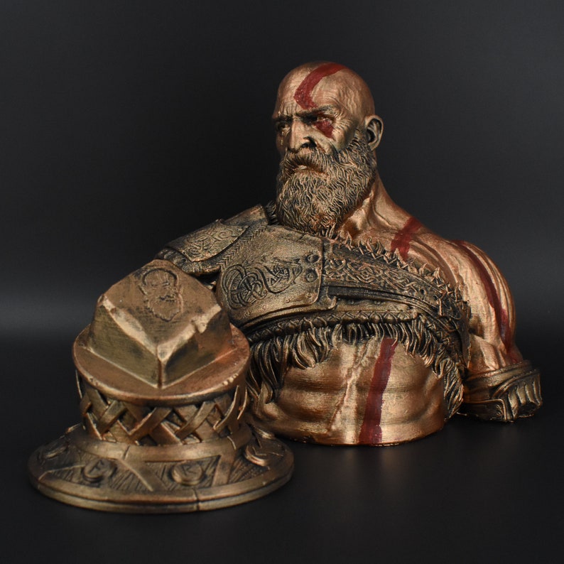 Puede incluir: Un busto de bronce de un guerrero con barba y marcas rojas en la cara. El busto lleva una armadura detallada con un dise&ntilde;o trenzado. El guerrero sostiene un escudo de bronce con un dise&ntilde;o en relieve.