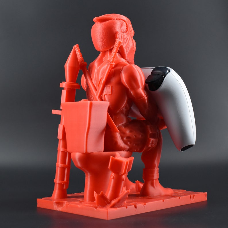 Peut inclure: Une figurine rouge en 3D imprim&eacute;e d'un personnage assis sur des toilettes tenant une manette de jeu vid&eacute;o blanche. La figurine est un personnage populaire d'un jeu vid&eacute;o.