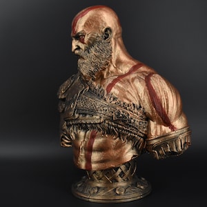 Puede incluir: Un busto de un personaje con barba y marcas rojas pintado de oro. El busto est&aacute; esculpido con una definici&oacute;n muscular detallada y una correa de cuero trenzada texturizada sobre el pecho.
