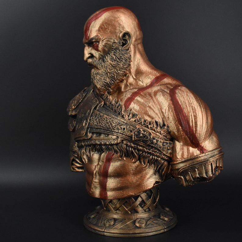 Puede incluir: Un busto de un personaje con barba y marcas rojas pintado de oro. El busto est&aacute; esculpido con una definici&oacute;n muscular detallada y una correa de cuero trenzada texturizada sobre el pecho.