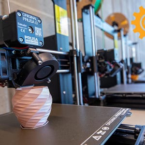 May include: A close-up of a 3D printer with a pink and white object being printed. The printer is a black and silver Original Prusa i3 model. The printer is in a workshop setting.