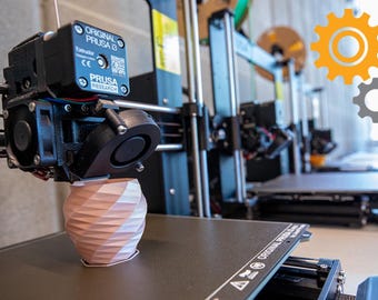 May include: A close-up of a 3D printer with a pink and white object being printed. The printer is a black and silver Original Prusa i3 model. The printer is in a workshop setting.