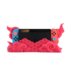 May include: A pink and blue Nintendo Switch console stand with a cloud design. The stand is made of plastic and has a cutout for the console's screen.