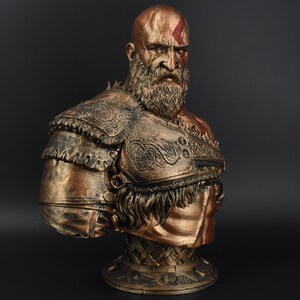 Puede incluir: Un busto de bronce de un guerrero calvo y barbudo con un dise&ntilde;o pintado en rojo y dorado en el pecho. El guerrero lleva una armadura detallada y adornada con flecos. El busto est&aacute; montado sobre una base con un patr&oacute;n tejido.