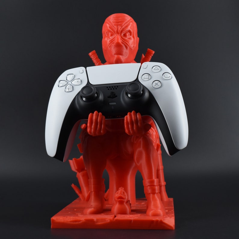 Peut inclure: Une figurine rouge en 3D d'un personnage tenant une manette PlayStation 5 DualSense blanche. La figurine est debout sur une base rouge.