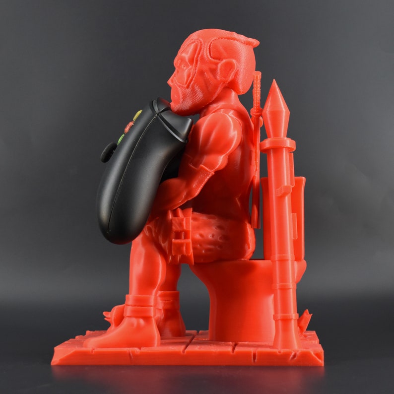 Peut inclure: Une figurine rouge en 3D imprim&eacute;e d'une personne assise sur des toilettes tenant une manette de jeu vid&eacute;o noire. La figurine tient la manette devant elle avec sa main gauche. La figurine porte une ceinture et a une &eacute;p&eacute;e sur le dos.