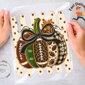 May include: A pumpkin-shaped graphic design featuring segments with football field markings, leopard print, brown footballs, and black and white buffalo plaid. A large black bow sits on top, set against a background of cream vertical stripes with brown dots.