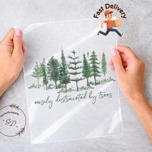 May include: A clear plastic sheet featuring a watercolor illustration of green trees. The text "easily distracted by trees" is written in a script font. A cartoon delivery person is running with a package.