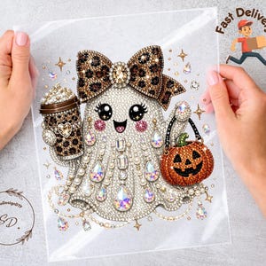 May include: A decorative transfer featuring a ghost character adorned with rhinestones, wearing a leopard print bow, holding a coffee cup with a leopard print sleeve, and carrying a small orange jack-o'-lantern bucket with a black handle.