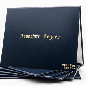 May include: A stack of navy blue diploma covers with gold lettering. The top cover is open, revealing a white interior. The text "Associate Degree" is printed in gold on the front. The bottom right corner of the cover has the text "Name Here" and "Date Here".