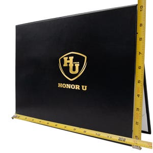 May include: A black certificate holder with a gold shield logo and the text HONOR U. A yellow measuring tape shows the holder is approximately 11.5 inches wide and 9 inches tall. The folder has a smooth finish and a rectangular shape.