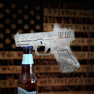Könnte beinhalten: Ein metallischer Flaschenöffner in Form einer Pistole mit silberner Oberfläche. Der Öffner wird verwendet, um eine braune Glasflasche Samuel Adams Bier zu öffnen. Der Flaschenöffner befindet sich vor einem unscharfen Bild einer amerikanischen Flagge.