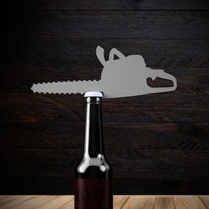 May include: A dark brown glass bottle of beer with a white cap and a label that says "BMF BEER" is balanced on top of a silhouette of a chainsaw.