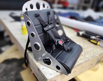 May include: A black racing seat with a silver metal frame and a black five-point harness. The seat has a red button on the harness.