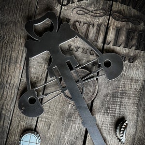 May include: A black metal bottle opener shaped like a crossbow. The opener is on a wooden surface with bottle caps scattered around it.