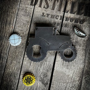 May include: A metal tractor bottle opener with a black finish. The bottle opener is on a wooden surface with the text "DISTILLER LYNCHBURG TENNESSEE" printed on the surface.