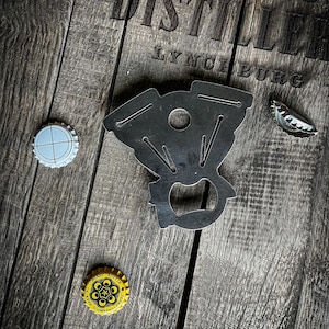 May include: A black metal bottle opener shaped like a car engine, with two bottle caps on a wooden surface. The surface has the text "DISTILLER LYNCHBURG" printed on it.