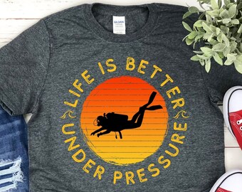 May include: A dark gray t-shirt with a graphic of a scuba diver in front of a sunset. The text "Life is better under pressure" is written in a circle around the diver.