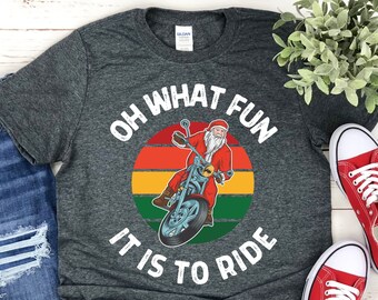 May include: A gray t-shirt with a white graphic of Santa Claus riding a motorcycle in front of a sunset. The text "Oh What Fun It Is To Ride" is written around the graphic.