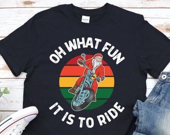 May include: Black t-shirt with a graphic of Santa Claus riding a motorcycle. The design includes the text "OH WHAT FUN IT IS TO RIDE" in a circular layout around the image of Santa and his bike. The background is a sunset design.