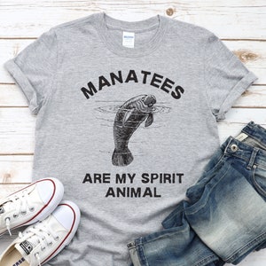 May include: A gray t-shirt with the words "MANATEES ARE MY SPIRIT ANIMAL" in black. The shirt features a black and white illustration of a manatee in water. White canvas sneakers and blue jeans are next to the shirt.