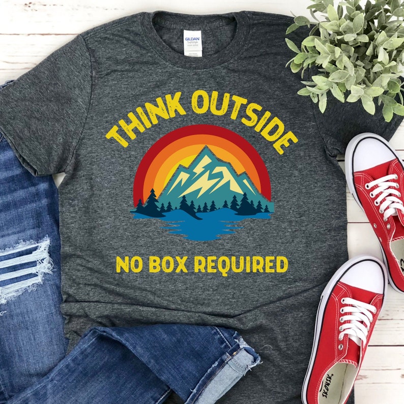 May include: A dark grey t-shirt with a retro-style mountain scene and the text "Think Outside No Box Required".