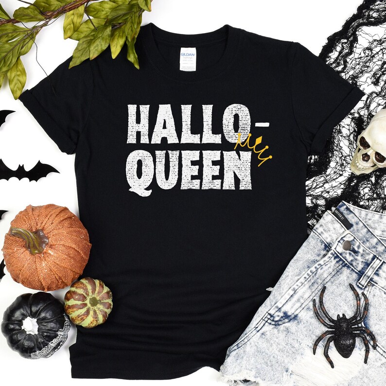 May include: Black t-shirt with white text that reads "Hallo-Queen" with a gold crown. The shirt is on a white background with pumpkins and a spider.