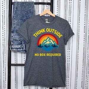 May include: A dark heather grey t-shirt with a retro mountain scene and the text "Think Outside No Box Required".