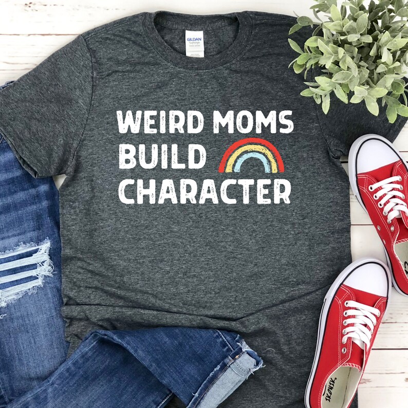 May include: A dark gray t-shirt with the text "Weird Moms Build Character" in white with a rainbow graphic. The shirt is laid out on a white wooden surface with blue jeans and red sneakers.