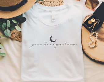 May include: A white t-shirt featuring a black crescent moon graphic and the text "your design here" in a cursive script. The shirt is displayed alongside a straw hat with a black band, brown sunglasses, and a small black jewelry box with pearls.