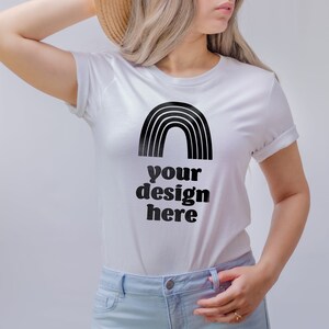 May include: A white short-sleeved t-shirt featuring a black graphic of a rainbow arch positioned above the bold black text that reads your design here. The shirt has a crew neckline and is paired with light blue denim jeans and a straw hat.
