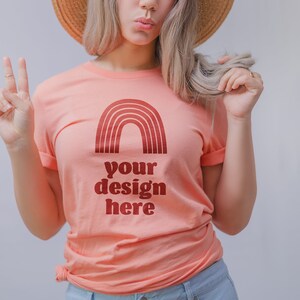 May include: A peach-coloured short-sleeve t-shirt featuring a dark red graphic of a rainbow arch above the text "your design here" printed in a bold, rounded font. The shirt has a crew neckline and is worn with a straw hat and light blue jeans.