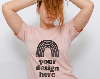 May include: A light pink short-sleeved t-shirt featuring a black graphic of a rainbow arch positioned above the bold black text that reads your design here. The garment has a crew neckline and is paired with light blue denim jeans.
